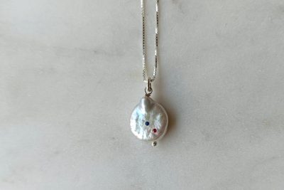 Μενταγιόν Disco Pearl με μαργαριτάρι σε χρώμα λευκό και σχήμα στρογγυλό επίπεδο & λεπτή βενετσιάνα αλυσίδα σε ασήμι 925. Στο μαργαριτάρι έχουν καρφωθεί ένα μπλε και ένα κόκκινο ζιργκόν. Μήκος αλυσίδας: 40 εκ.